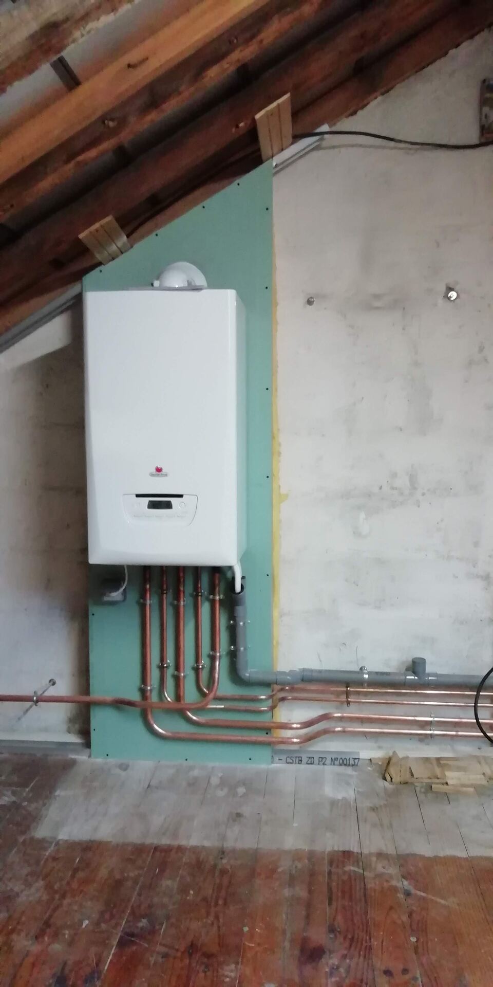 Chaudière murale à gaz fixée sur un panneau vert sous les rampants d'un comble, avec un collecteur de tubes cuivre cintrés à sa base.