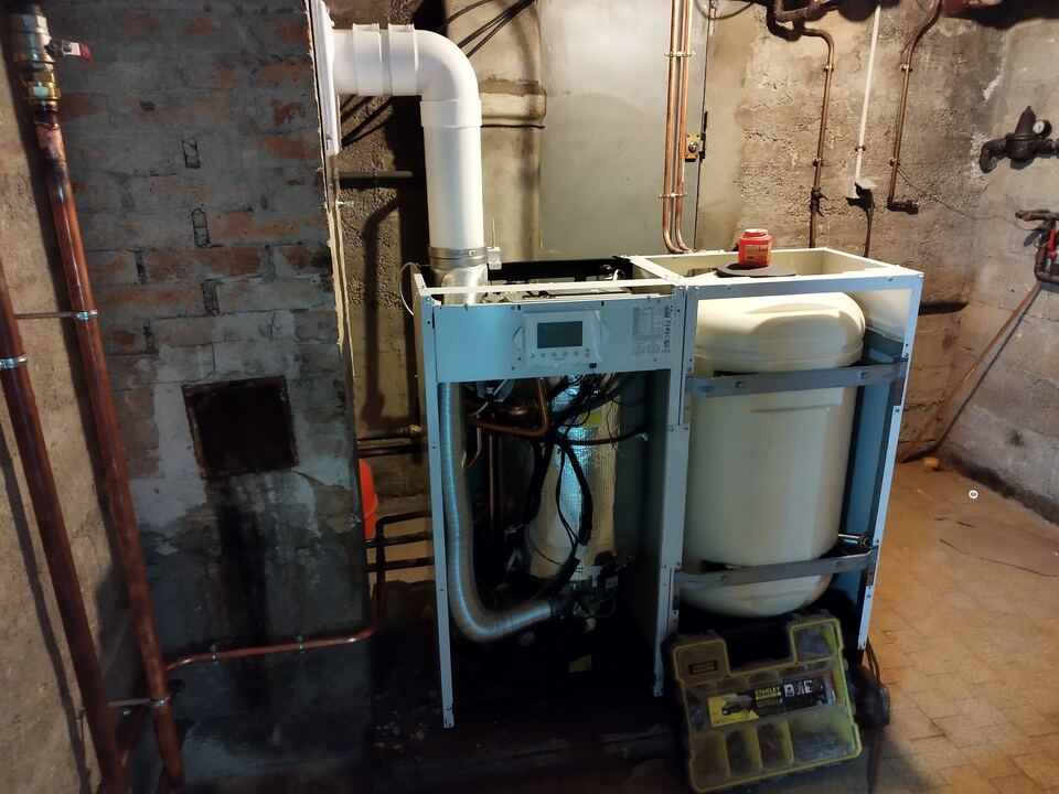 Chaudière capot ouvert en cours de raccordement dans une cave aux murs de pierre et de brique, à côté d'un ballon d'eau chaude.